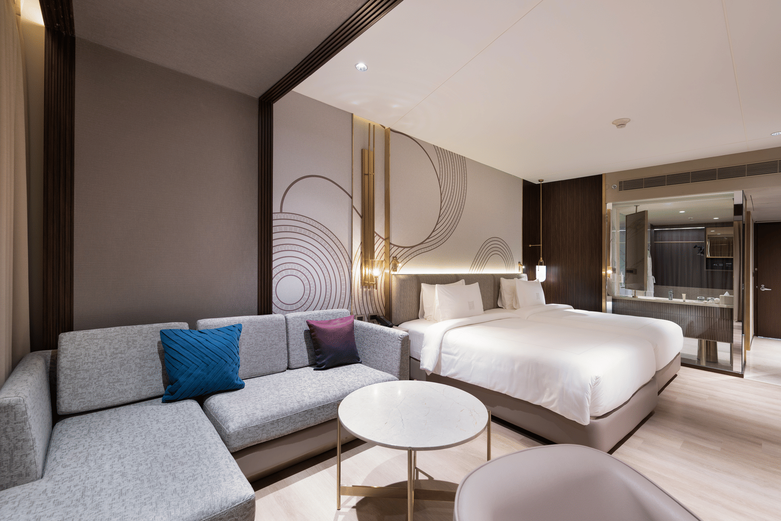 A wide shot of a hotel suite showing a large bed, a modern sofa seating area, and tasteful decor at Merlis Hotel, Coimbatore.