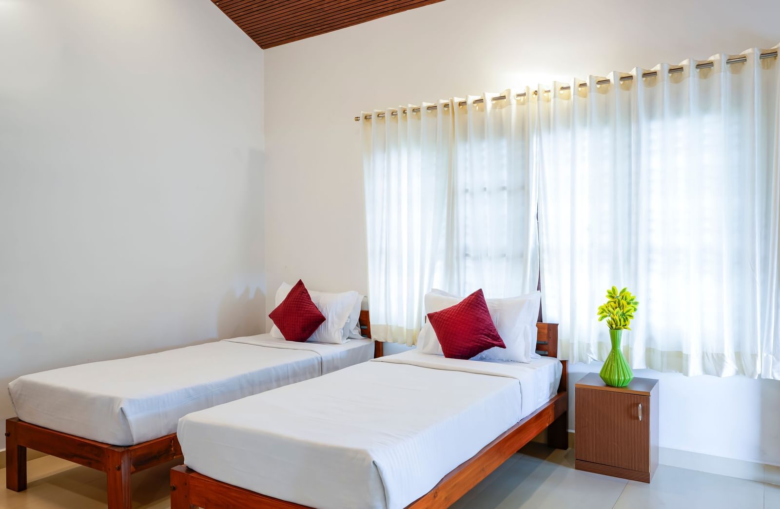 A bright hall of residences at Bhuthanakadu Retreat, featuring two single beds with crisp white bedding and red accent pillows, and large windows with natural light.
