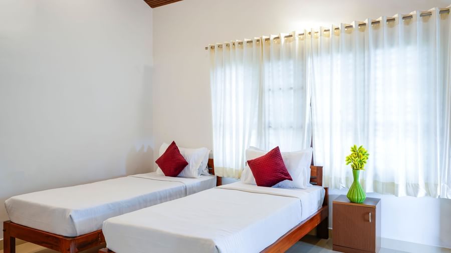 A bright hall of residences at Bhuthanakadu Retreat, featuring two single beds with crisp white bedding and red accent pillows, and large windows with natural light.