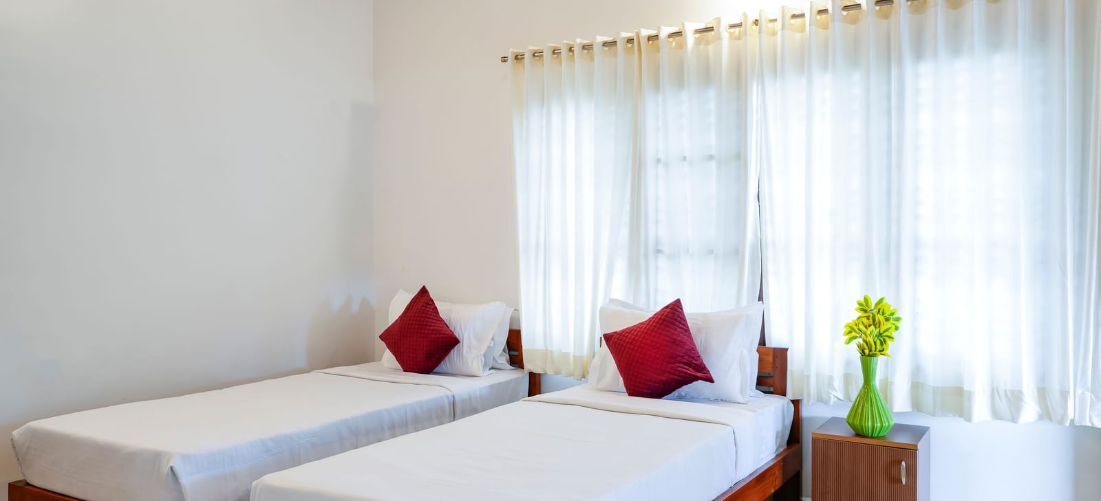 A bright hall of residences at Bhuthanakadu Retreat, featuring two single beds with crisp white bedding and red accent pillows, and large windows with natural light.