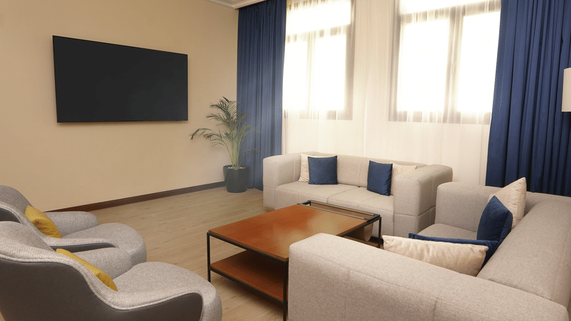 A corner view of the deluxe two bedroom apartment living area at La Maison, Doha, with a couch, chairs, a coffee table, and a television on the wall.