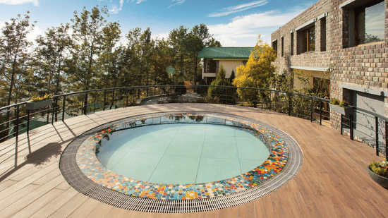 Circular outdoor pool with colourful mosaic tiles around the edge, set on a wooden deck with a metal railing, surrounded by trees and modern stone buildings under a clear blue sky - Suryavilas Luxury Resort and Spa