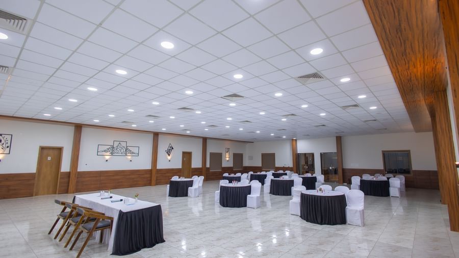The spacious banquet hall at the Daksh Eden Greenz with circular tables and chairs placed in a banquet style along with overhead lighting.