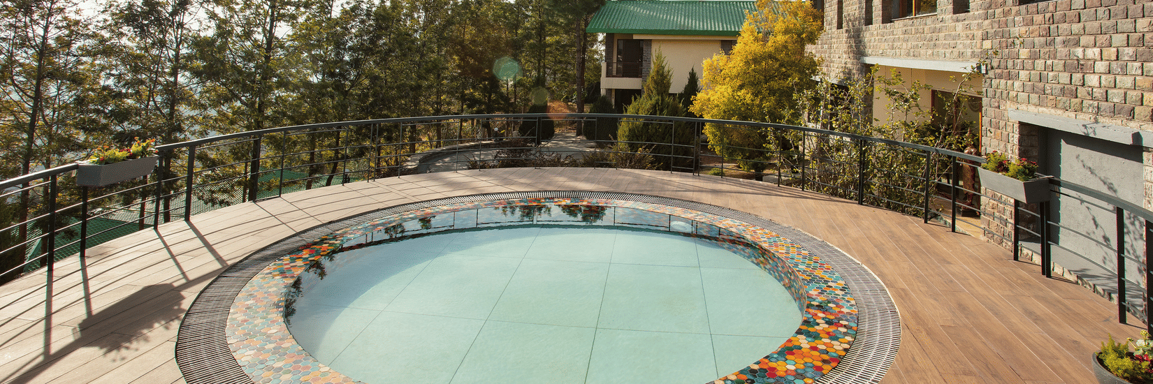 Circular outdoor pool with colourful mosaic tiles around the edge, set on a wooden deck with a metal railing, surrounded by trees and modern stone buildings under a clear blue sky - Suryavilas Luxury Resort and Spa