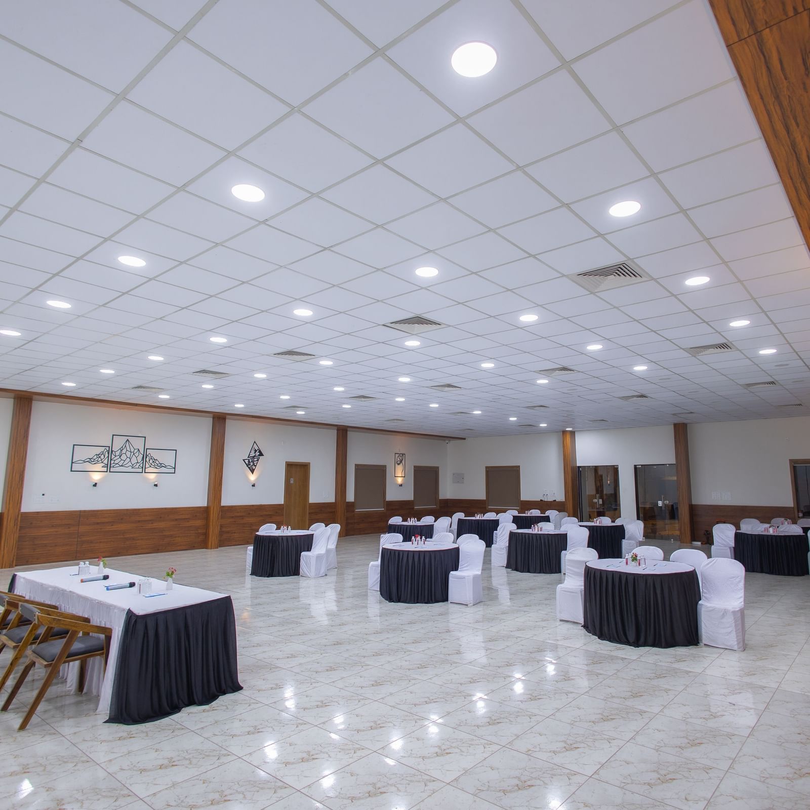 The spacious banquet hall at the Daksh Eden Greenz with circular tables and chairs placed in a banquet style along with overhead lighting.