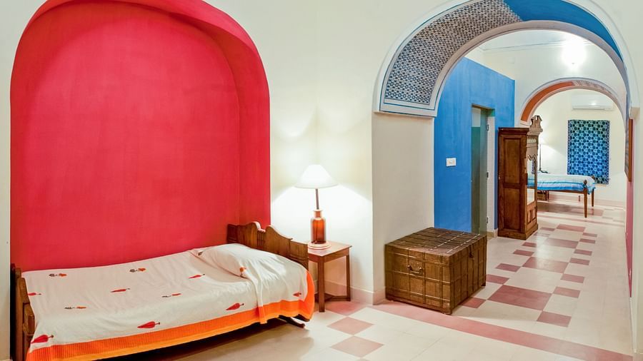 Hotel room with a single bed in a red alcove, a bedside table with a lamp, a chest, and a view through an archway to another room with a bed at Tijara Fort-Palace, Alwar.