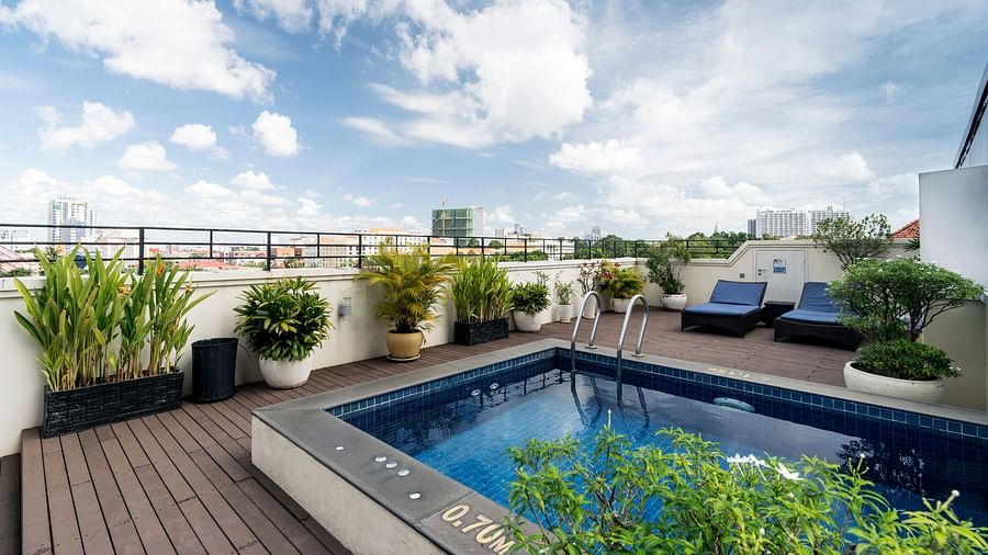 A rooftop terrace with a small pool or hot tub is visible, surrounded by potted plants and a railing, under a bright sky with scattered clouds | Central Mansions, Phnom Penh