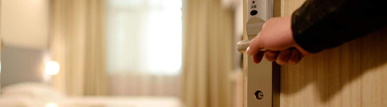 A hand opening a hotel room door, revealing a modern, well-lit room with a neatly made bed, a small round table, and curtains.