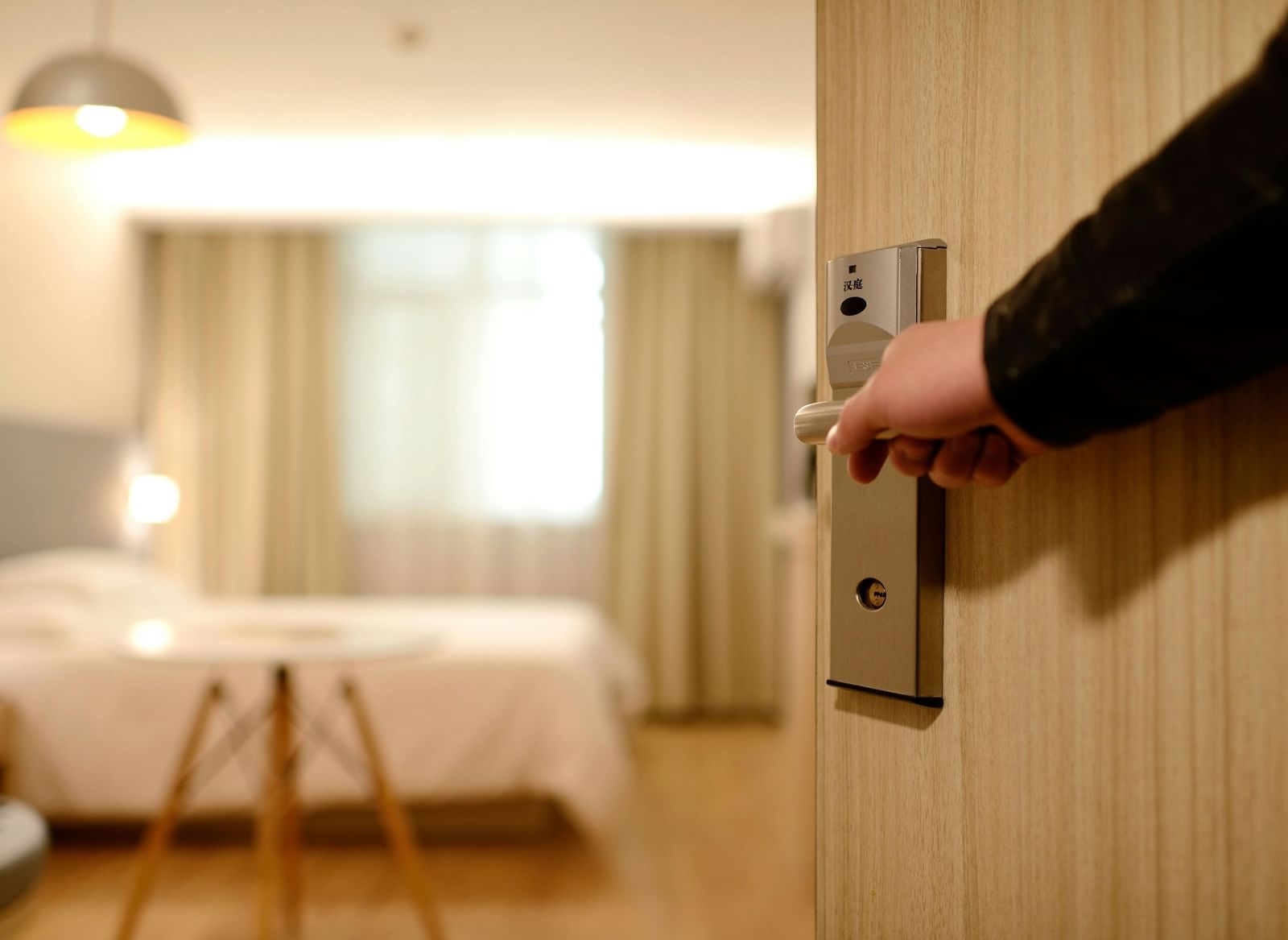 A hand opening a hotel room door, revealing a modern, well-lit room with a neatly made bed, a small round table, and curtains.