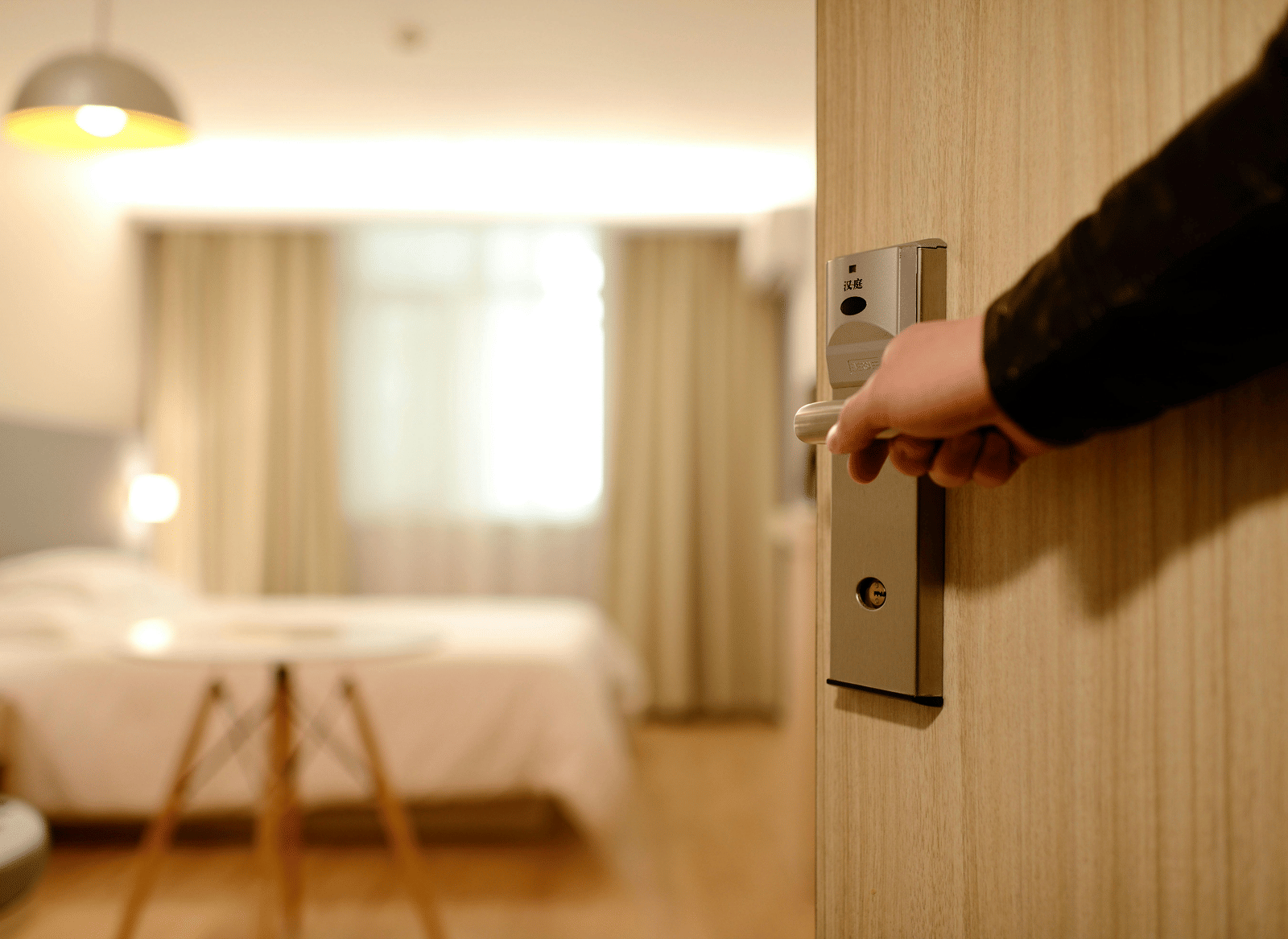 A hand is pushing open a light wood door, revealing a blurred, well-lit hotel room inside.