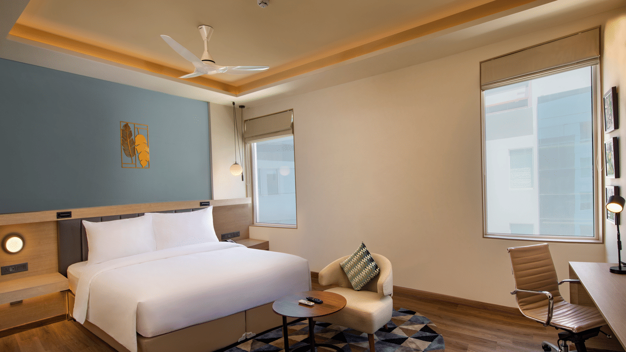 An Elite King Room at O By Tamara, Coimbatore, featuring a well-made bed, a workspace with a desk and a chair, a ceiling fan, and 2 windows.