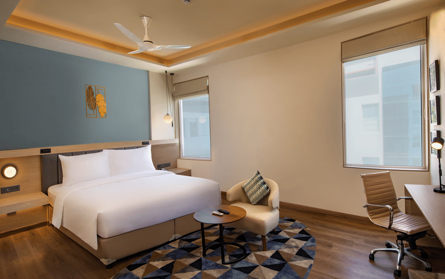 An Elite King Room at O By Tamara, Coimbatore, featuring a well-made bed, a workspace with a desk and a chair, a ceiling fan, and 2 windows.