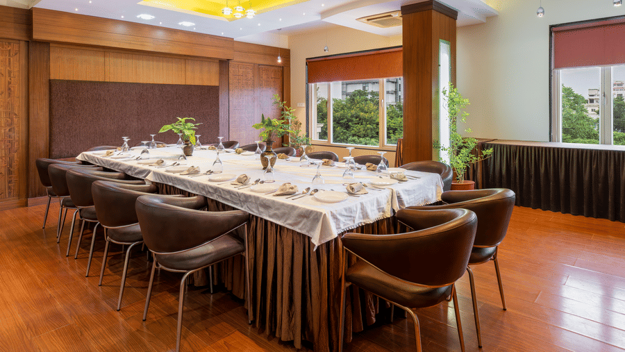 A professional boardroom setup featuring a long conference table with brown leather chairs, elegant wooden wall panelling, with ample of natural light coming from the windows - Amantra Comfort Hotel