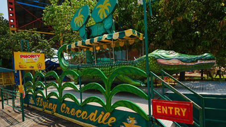 Crocodile ride with a green crocodile-shaped car, striped awning and palm-leaf fans, Flying Crocodile, Priority Line and Entry signs at MGM Dizzee World, Chennai.