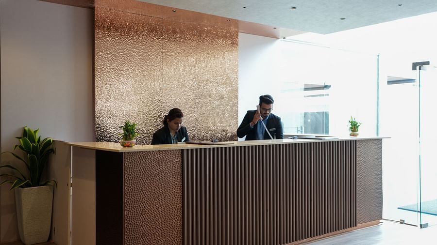 Two receptionists at a wooden front desk, one on the phone and the other working on a computer at TGI Livancia Hillside, JC Nagar, Mysore