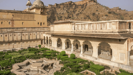 View of a geometric sunken garden and arched stone corridors within the Amer Fort complex, with hills in the distance.