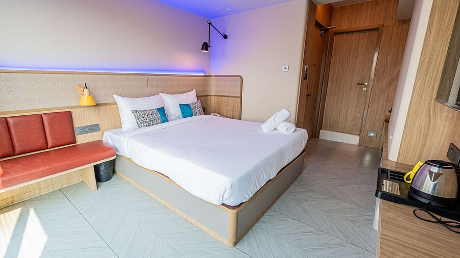 A spacious room at Indie Stays Mumbai International Airport, featuring cushioned furniture beside the bed and a wooden doorway leading to another room.