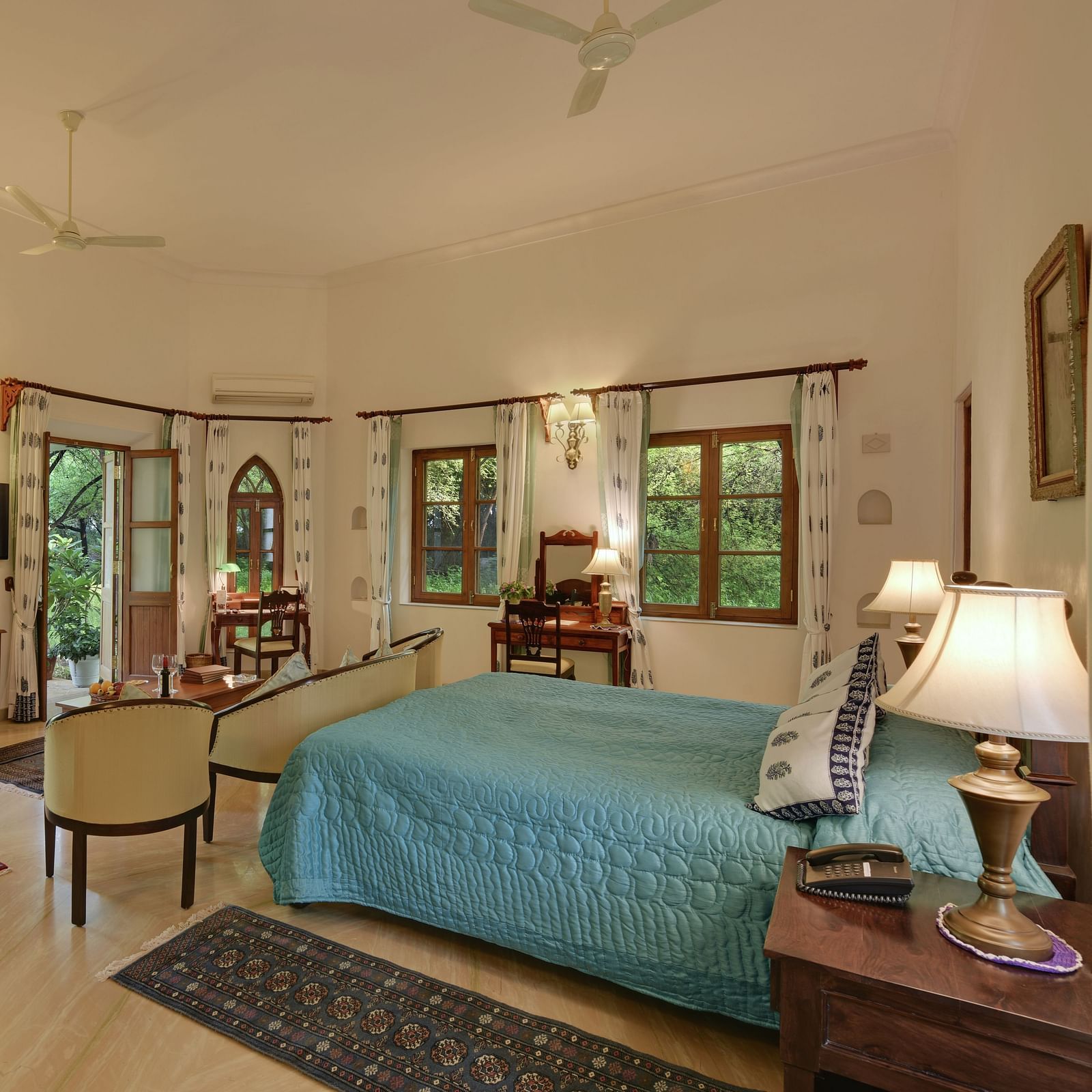 A bright, spacious room with a made bed, seating area, and multiple windows leading to an outdoor space, featuring a polished floor- Shahpura Bagh.
