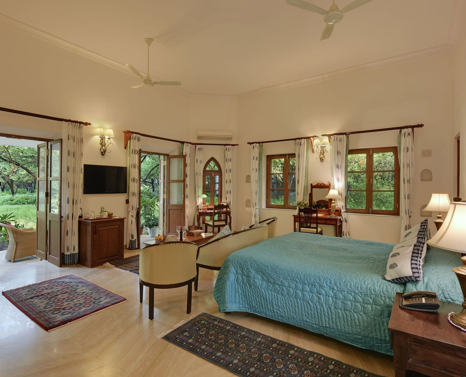 A bright, spacious room with a made bed, seating area, and multiple windows leading to an outdoor space, featuring a polished floor- Shahpura Bagh.