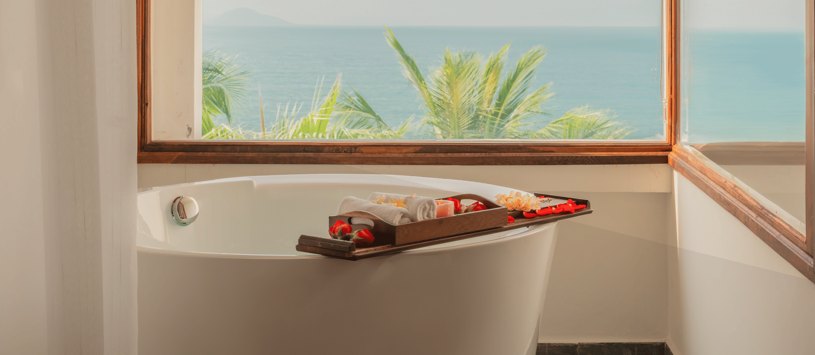 Freestanding bathtub by window at Alibu Resort Nha Trang, Vietnam, overlooking palm trees and sea with spa tray, wooden surround and tiled floor.