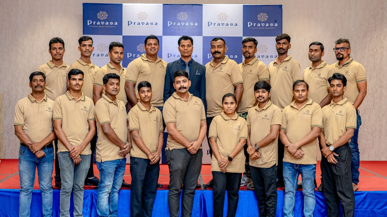 A large group of men in matching tan shirts and blue jeans stand in rows in front of a blue and white backdrop.