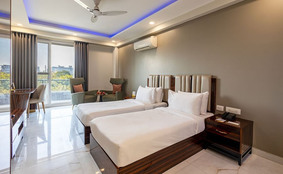 A room with twin beds, soft lighting, a ceiling fan, and a huge glass door at Hotel Bawa, Gurgaon.