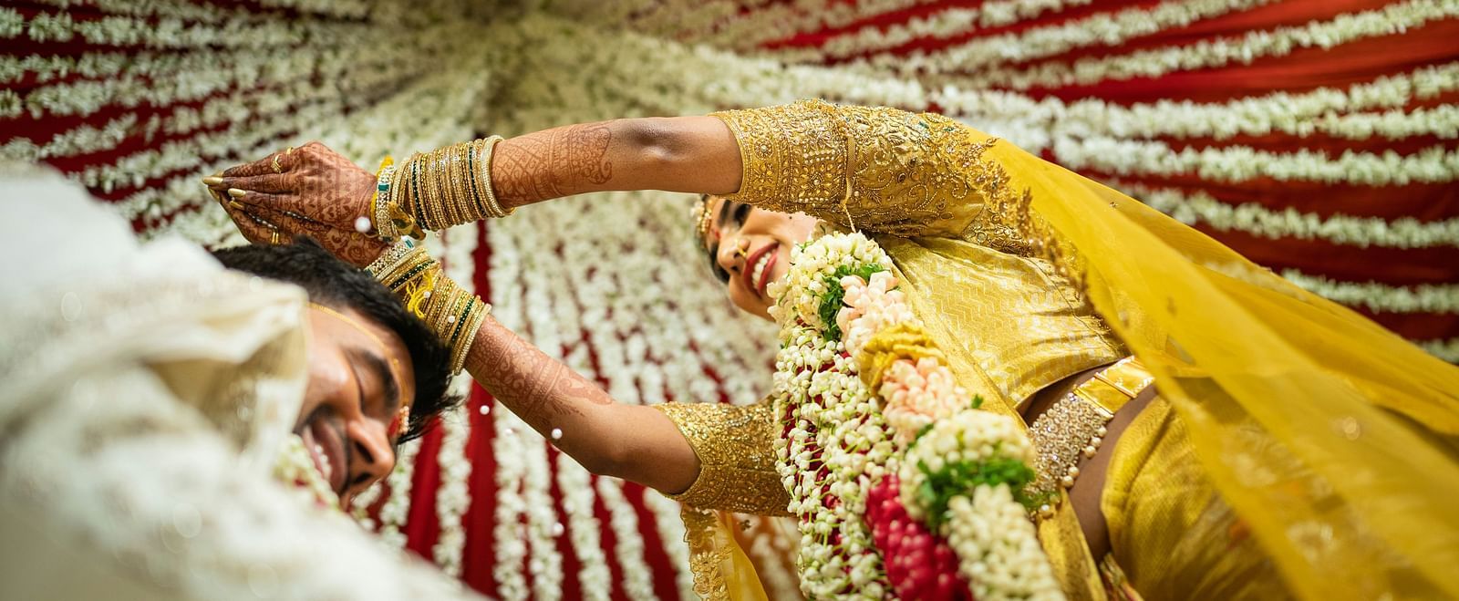A lower angle shot of a bride adored with a flower garland dressed in a yellow dress standing in front of the broom.