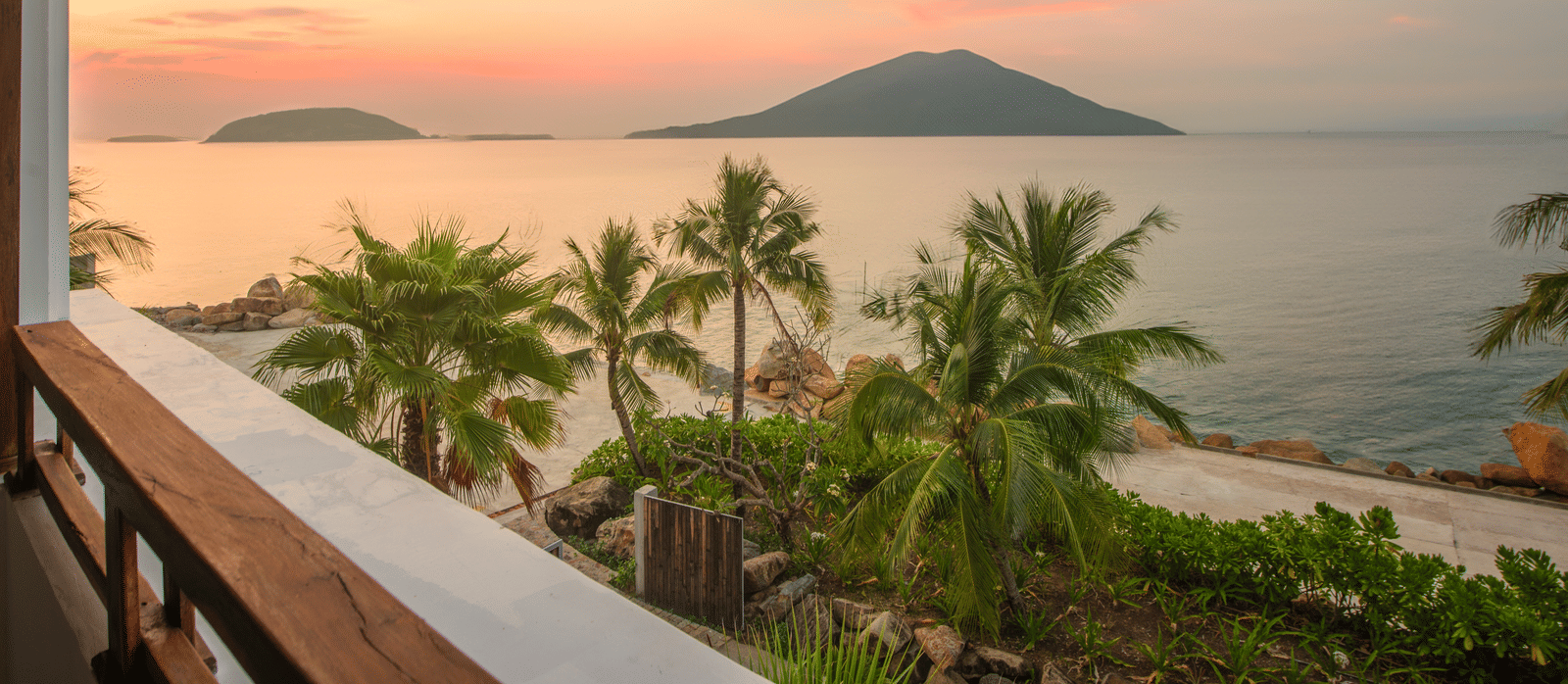 Sunset view from Alibu Resort Nha Trang, Vietnam, showing balcony, trees, island silhouette, pink sky and calm sea.