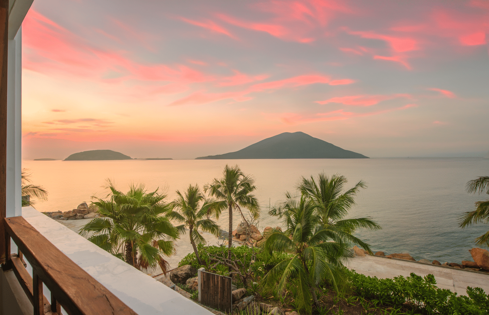 Sunset view from Alibu Resort Nha Trang, Vietnam, showing balcony, trees, island silhouette, pink sky and calm sea.