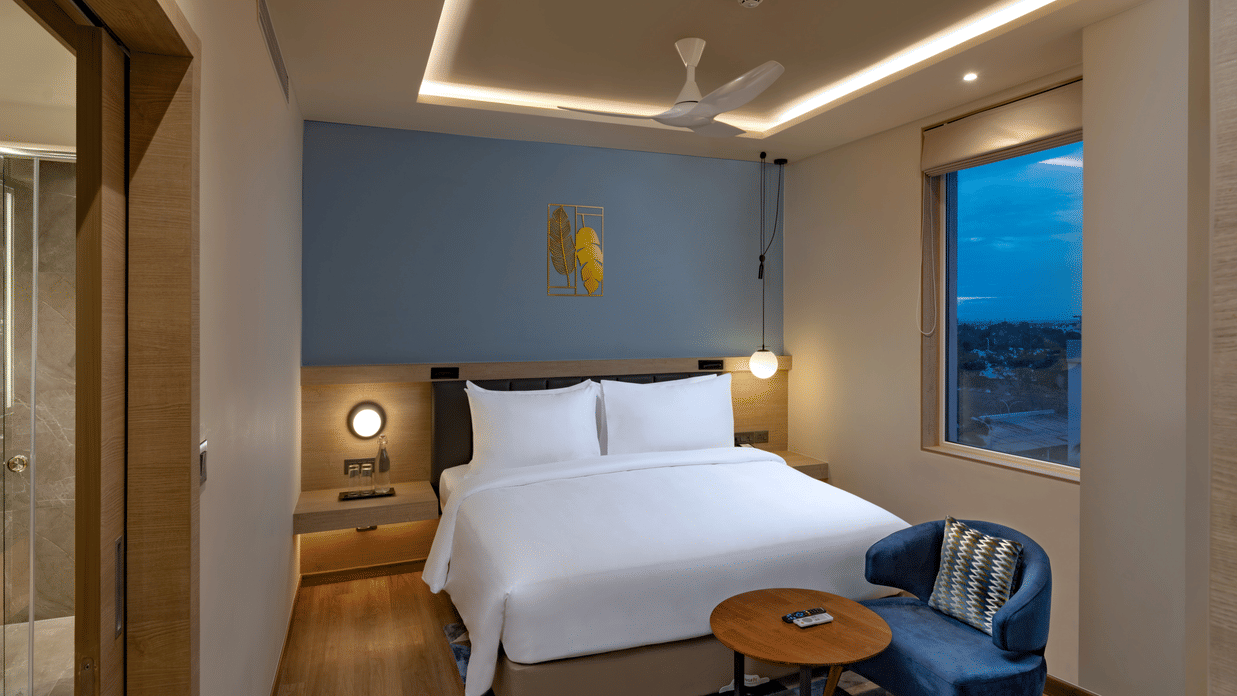 An Elite King Room at O By Tamara, Coimbatore, featuring a neatly made bed, a chair facing a small table, wooden flooring, and a window offering the view outside. 
