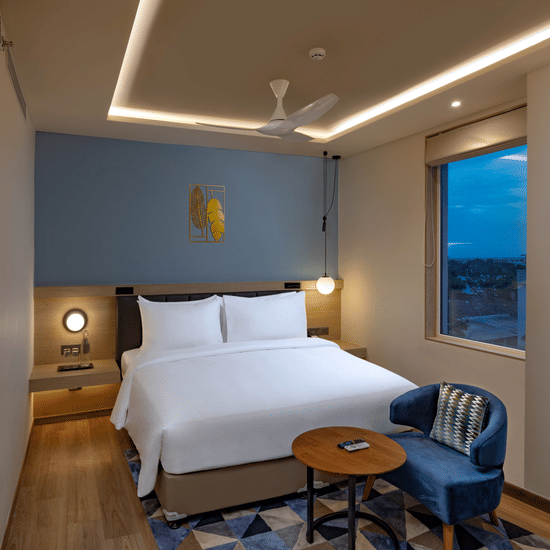 An Elite King Room at O By Tamara, Coimbatore, featuring a neatly made bed, a chair facing a small table, wooden flooring, and a window offering the view outside. 