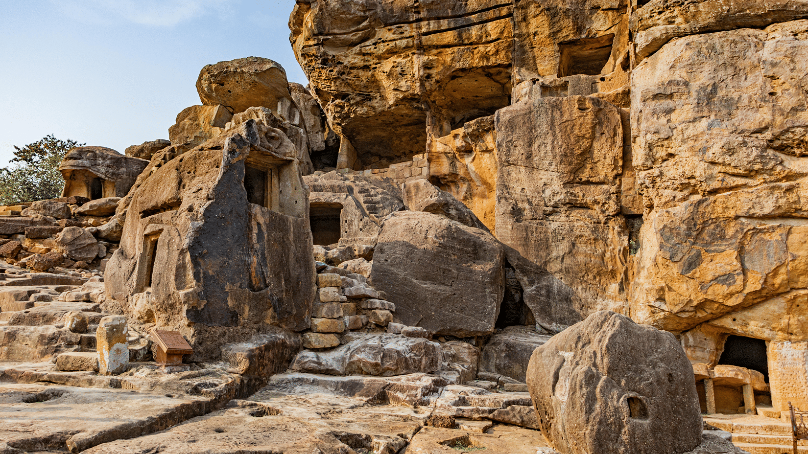 Ancient sandstone caves and rugged rock formations with carved steps, showcasing a weathered archaeological site beneath a clear blue sky