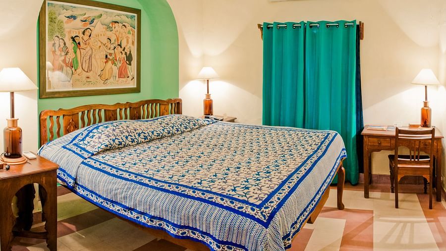Bedroom with a bed featuring blue and white patterned bedding, bedside tables with lamps, a desk, artwork in a green alcove, teal curtains, and AC at Neemrana's Tijara Fort Palace, Alwar.