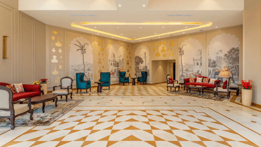 A spacious lobby lounge at EsthereaRaj Leela, Ranakpur, with red and teal armchairs, tall windows, and wall murals of palm trees.