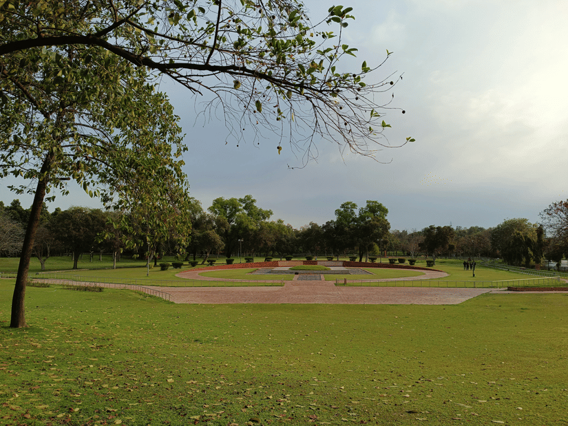 A wide view of a large, grassy public park with trees, a central paved or coloured area, and an overcast sky.
