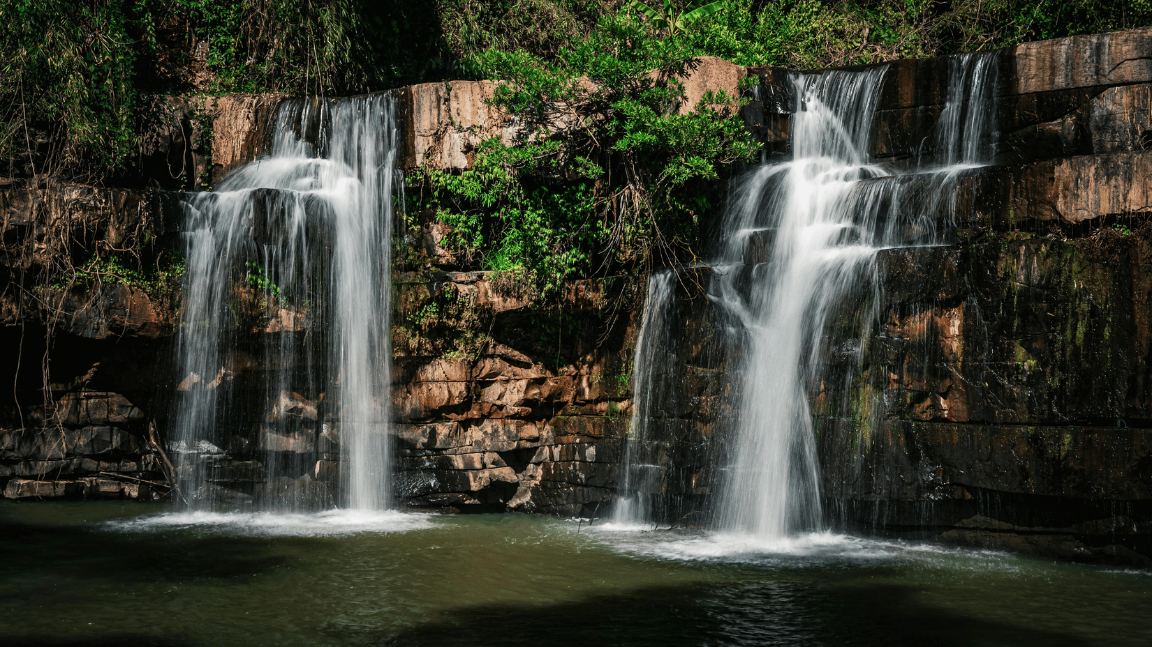 Two side-by-side waterfalls flowing over a rocky ledge into a dark pool of water below.