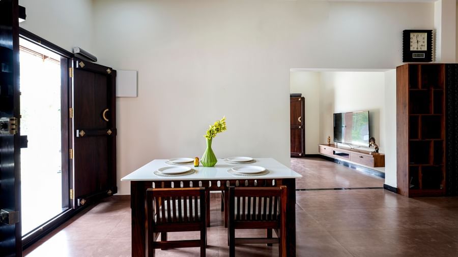 The dining area of the villa at Bhuthanakadu Retreat, featuring a simple wooden dining table and chairs set with a vase of flowers, next to large glass doors leading to the outside.