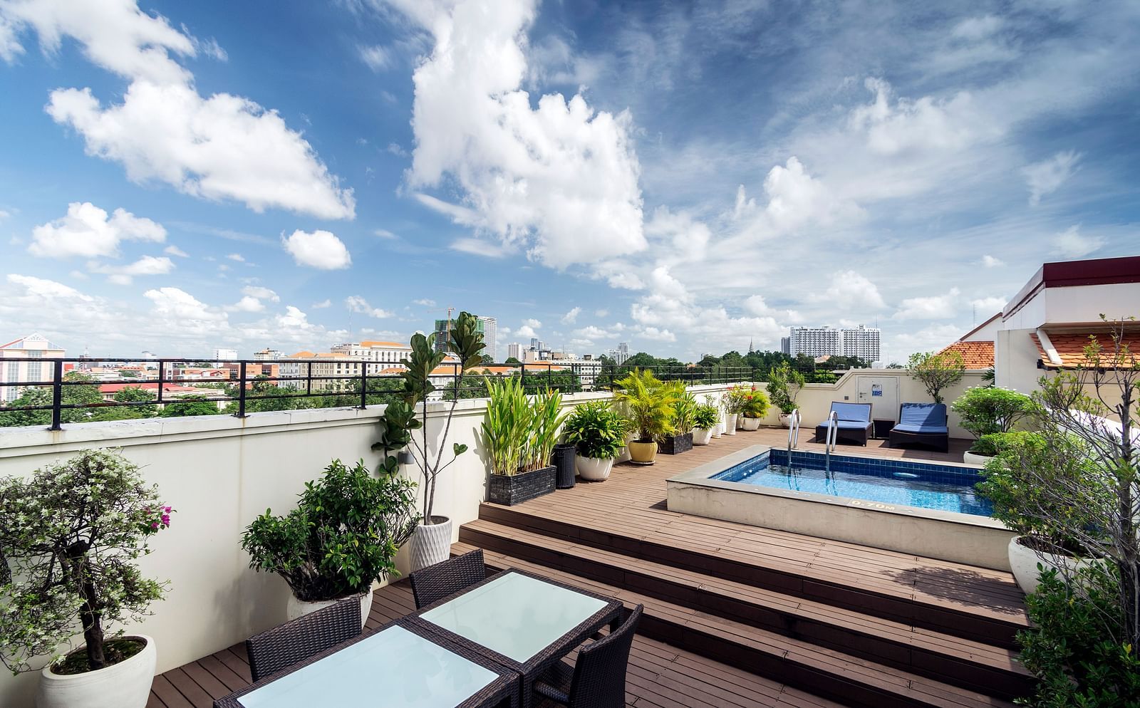 A rooftop terrace features a swimming pool, lush greenery, and comfortable seating under a clear blue sky with white clouds | Central Mansions, Phnom Penh