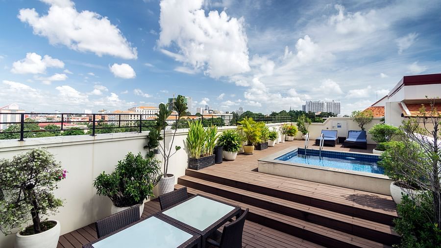 A rooftop terrace features a swimming pool, lush greenery, and comfortable seating under a clear blue sky with white clouds | Central Mansions, Phnom Penh