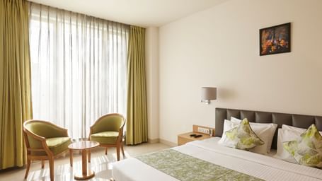 A bright hotel room at Monday Hotels Mahape, Navi Mumbai, with a bed, seating chairs by the window, and a TV.