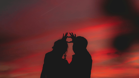 A view from below of a couple's silhouette making a heart shape with their hands with different hues in the sky.