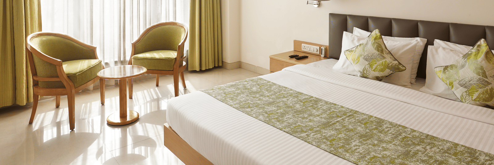 A bright hotel room at Monday Hotels Mahape, Navi Mumbai, with a bed, seating chairs by the window, and a TV.
