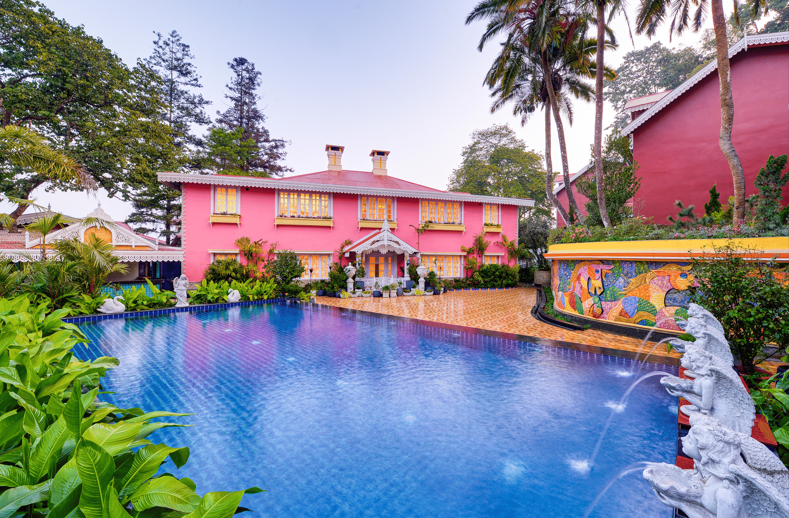 Outdoor pool on the deck beside a pink heritage building, with a mosaic mural, fountains and tropical plants at MAYFAIR Himalayan Spa Resort, Kalimpong.