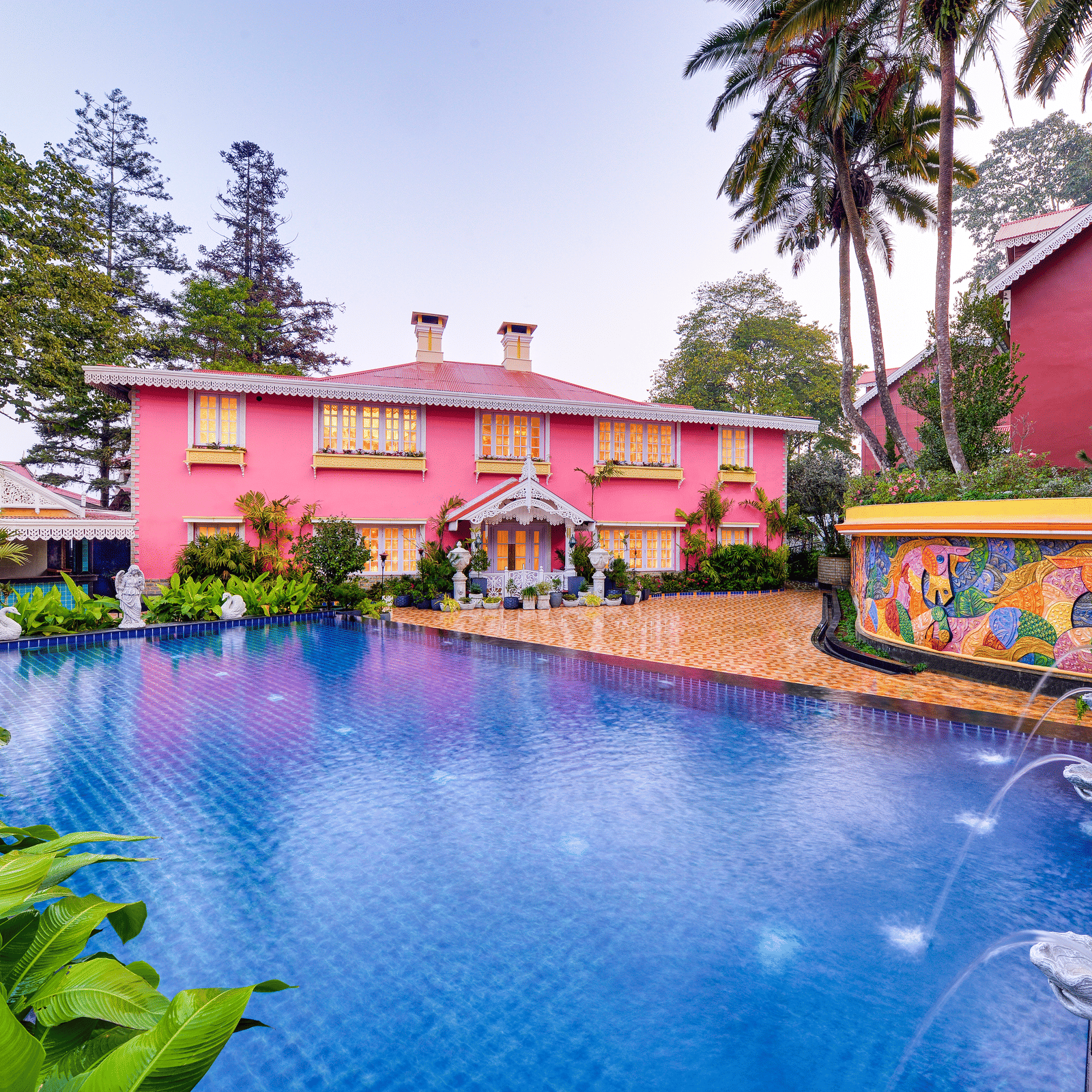Outdoor pool on the deck beside a pink heritage building, with a mosaic mural, fountains and tropical plants at MAYFAIR Himalayan Spa Resort, Kalimpong.