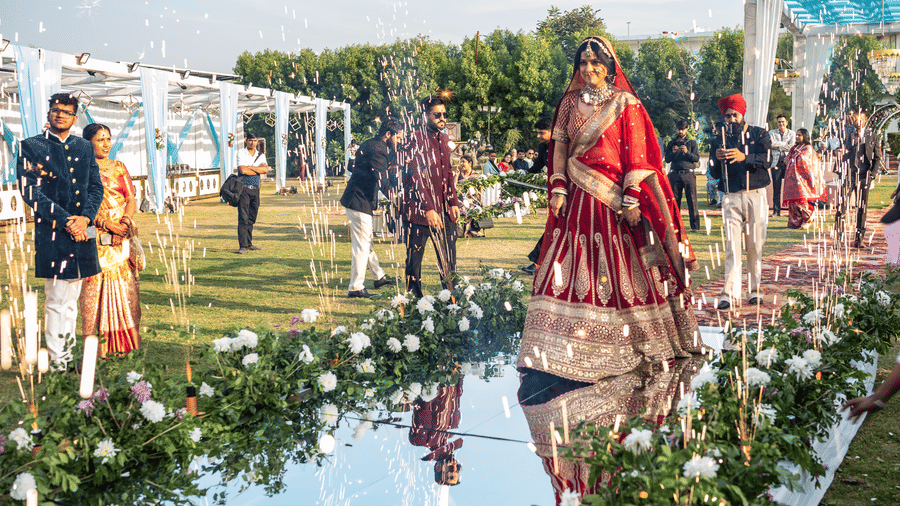 A vibrant shot of wedding guests, including a bride in a red lehenga, walking on a reflective floor decorated with white flowers at an outdoor function at Vedikant Resorts The Mallard Corbett.