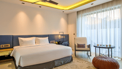 A plush bed and a sitting space by the large window consisting of a chair next to a coffee table and a pouffe at Azaya Beach Resort Goa