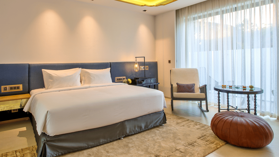 A plush bed and a sitting space by the large window consisting of a chair next to a coffee table and a pouffe at Azaya Beach Resort Goa