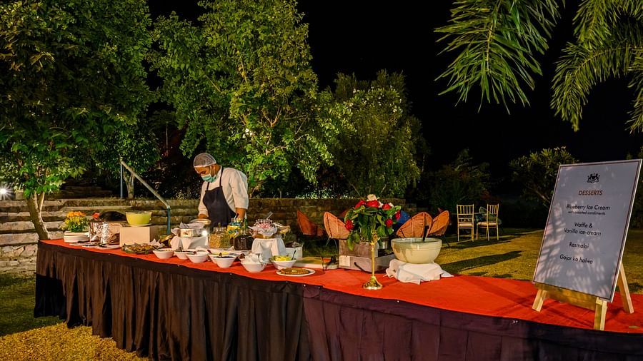 An outdoor buffet with various dishes and a person serving is set up at night at Tijara Fort-Palace - 19th Century, Alwar, surrounded by trees.