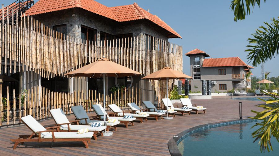 Row of wooden sun loungers with white cushions and parasols on a wooden deck next to a swimming pool, and a stone building at Hotel Sonar Bangla, Sundarban.