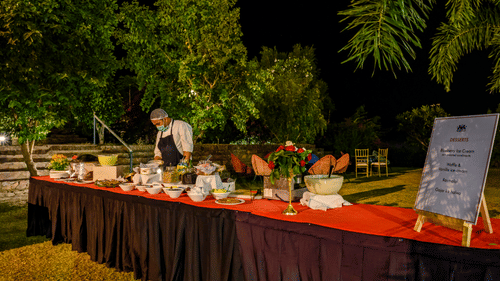 An outdoor buffet with various dishes and a person serving is set up at night at Tijara Fort-Palace - 19th Century, Alwar, surrounded by trees.